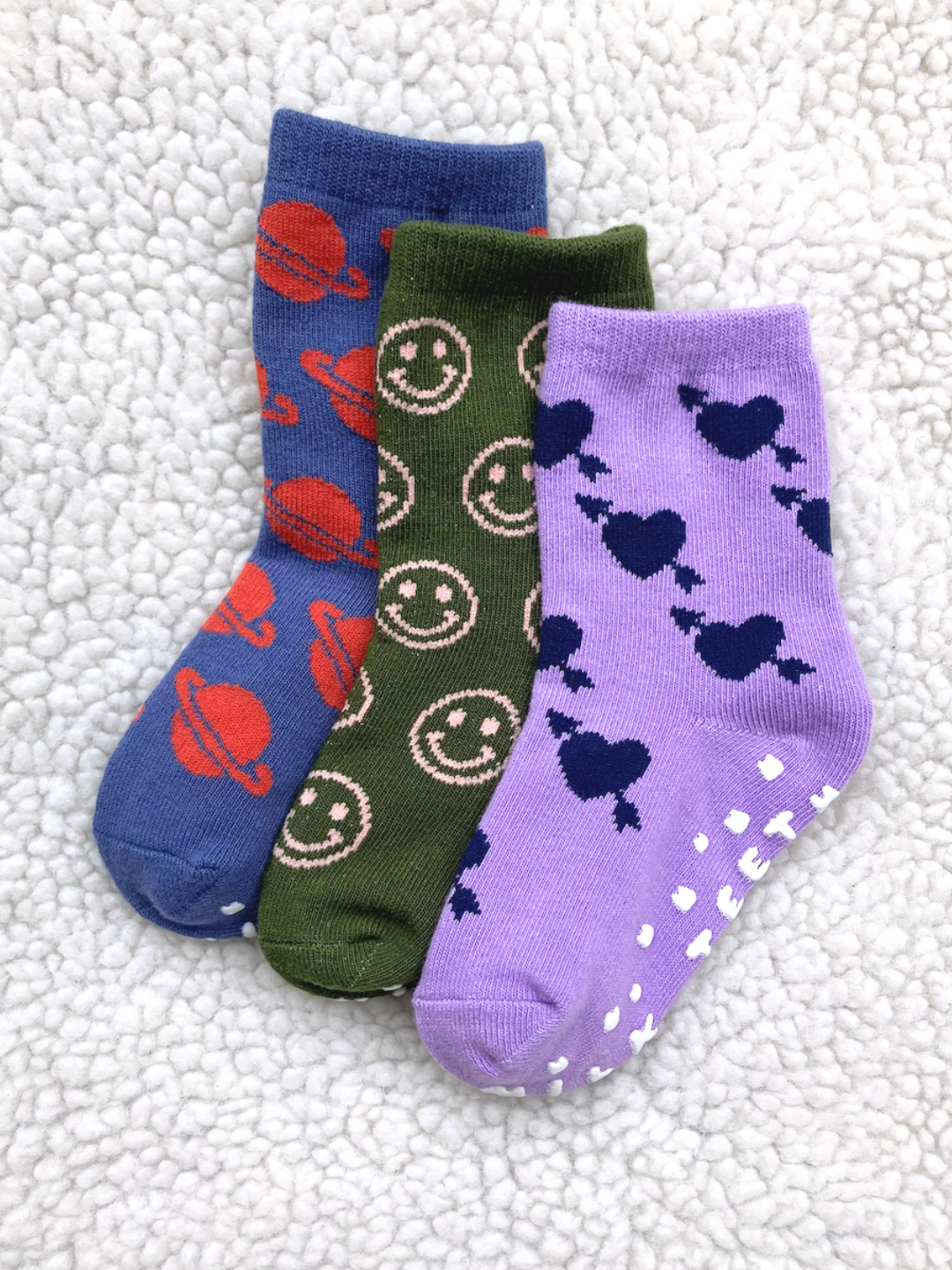 Olive Smile | Three pairs of GRIP SOCK children's socks on a fluffy white surface; designs include stretchy blue socks with red planets, green socks with tan smiley faces, and purple anti-skid socks with dark blue hearts.