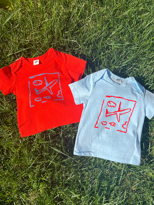 Second image of Light Blue | The AIRPLANE BABY TEE is a light blue short-sleeve toddler shirt with a playful red abstract airplane and cloud print on the front, shown against a plain white background.