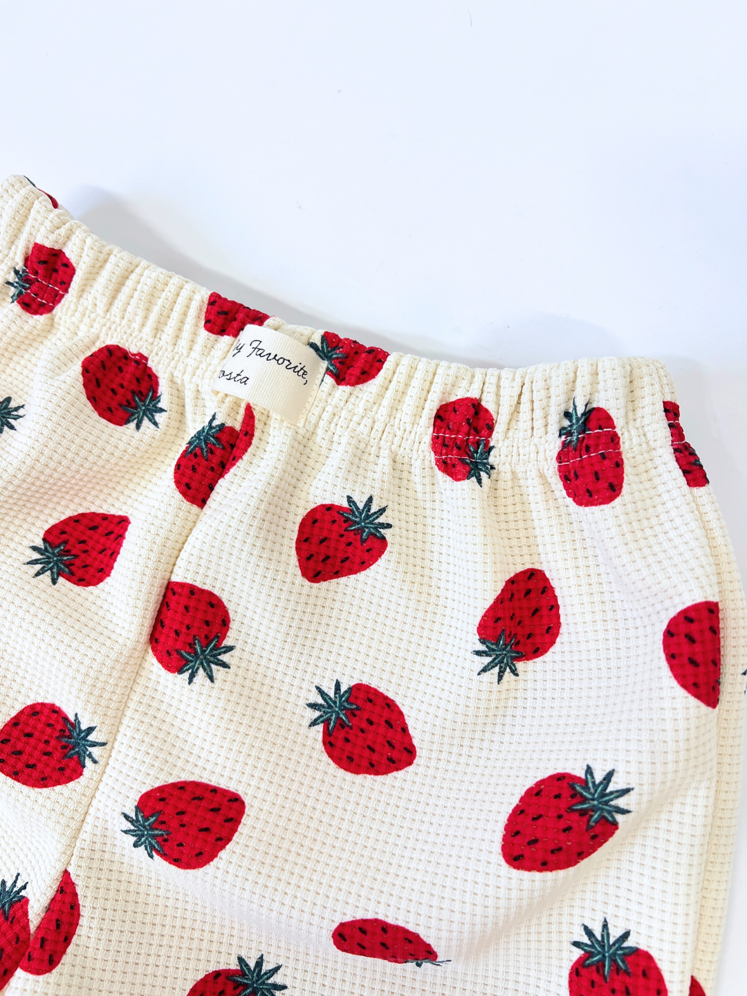 The STRAWBERRY WAFFLE SHORTS feature a patterned fabric of red strawberries on a white background, with the waistband including a small tag text.