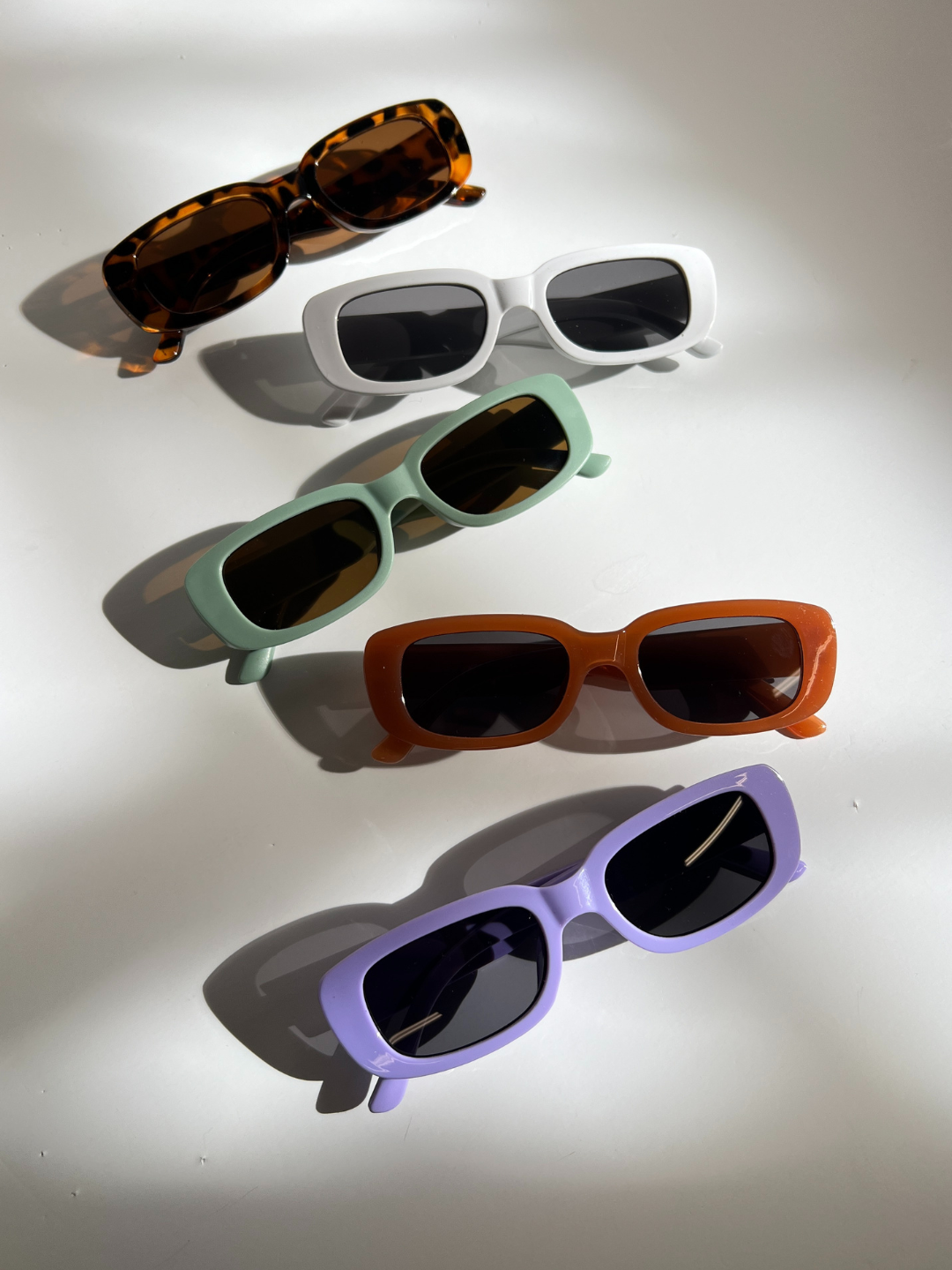 Lavender | Five pairs of SANTA CRUZ SUNGLASSES with UV400 lenses feature tortoiseshell, white, green, brown, and purple frames. Arranged diagonally on white, these stylish sunglasses protect the eyes of fashion-forward kids aged 2-6.