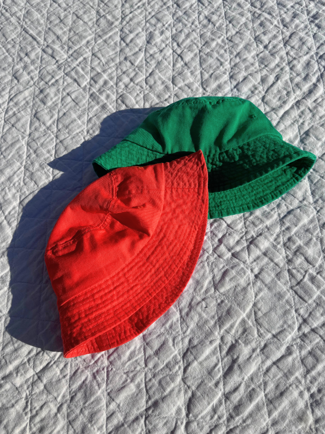 TWO KID'S PICNIC BUCKET HATS  IN TOMATO ORANGE AND KELLY GREEN ARE LAID FLAT ON BLANKET