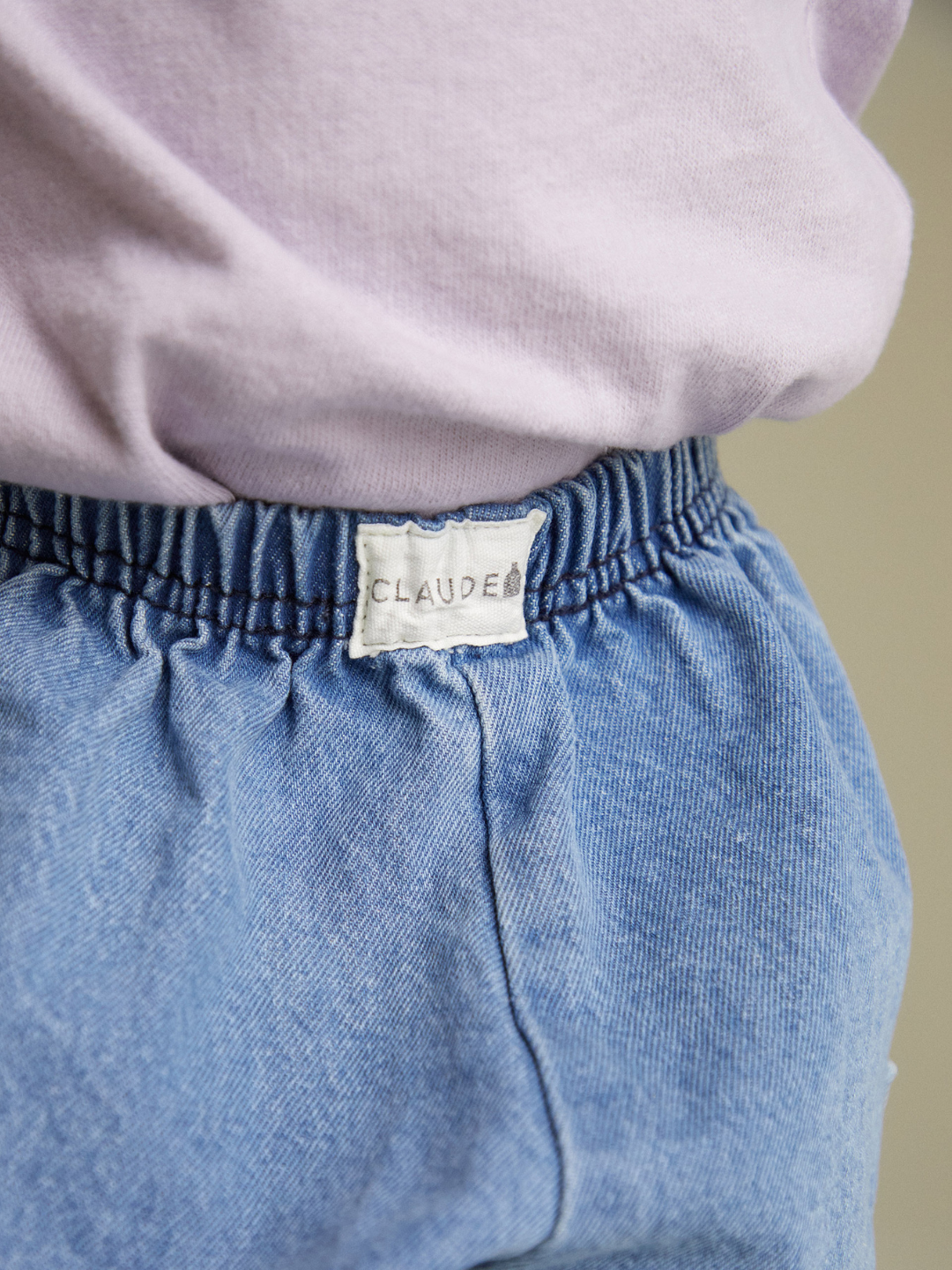 Close-up of a person wearing EMERSON DENIM BABY TROUSER in light blue organic cotton with an elastic waist, paired with a light purple top; a label reading "CLAUDE" is visible on the pants.