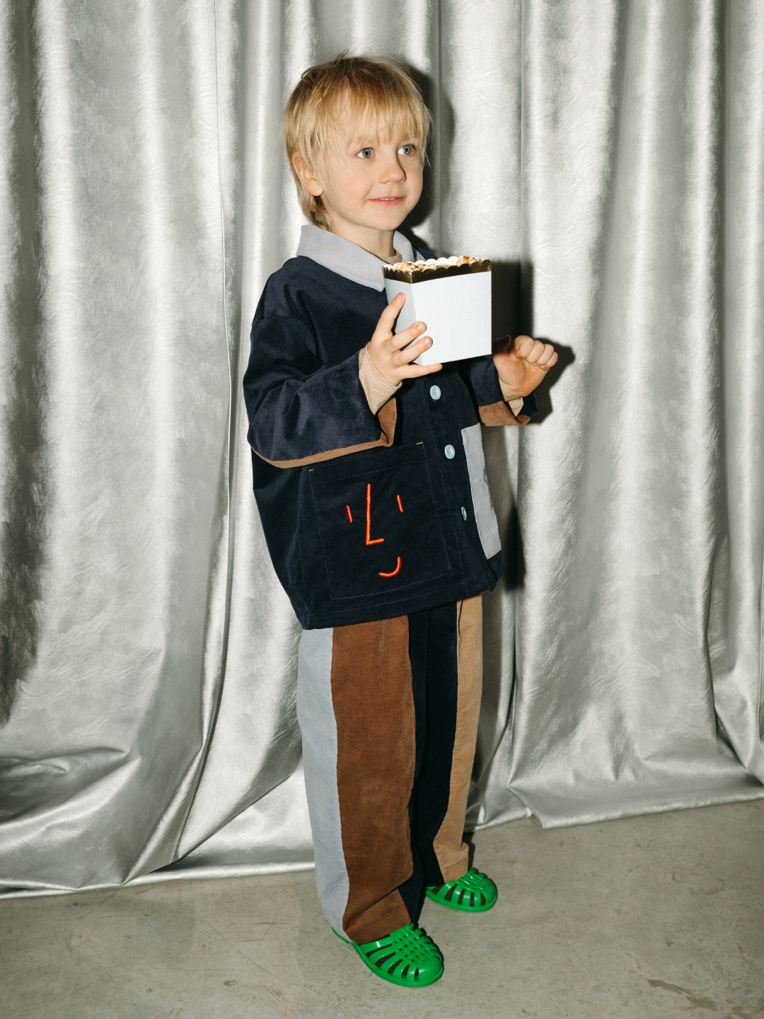 A young child wearing the FACE JACKET and green sandals stands on a concrete floor in front of silver curtains, holding a box of popcorn.