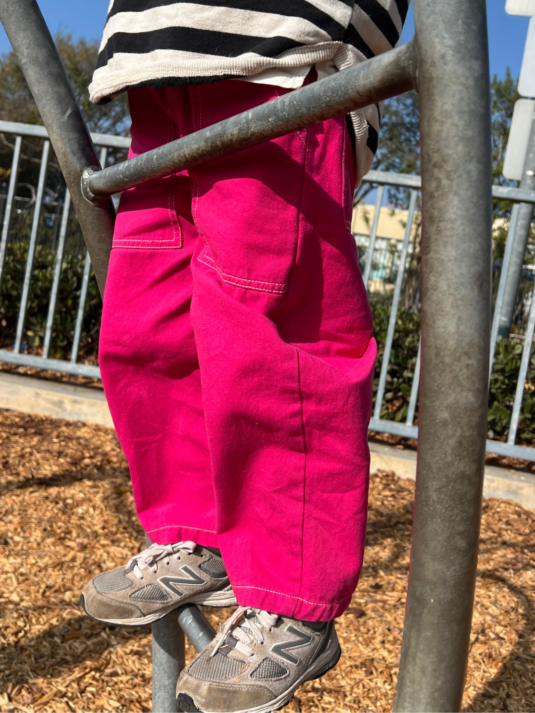 A child is joyfully climbing the playground's metal ladder wearing relaxed pull-on TOPSTITCH TWILL PANTS in bright pink soft cotton twill, paired effortlessly with gray shoes.