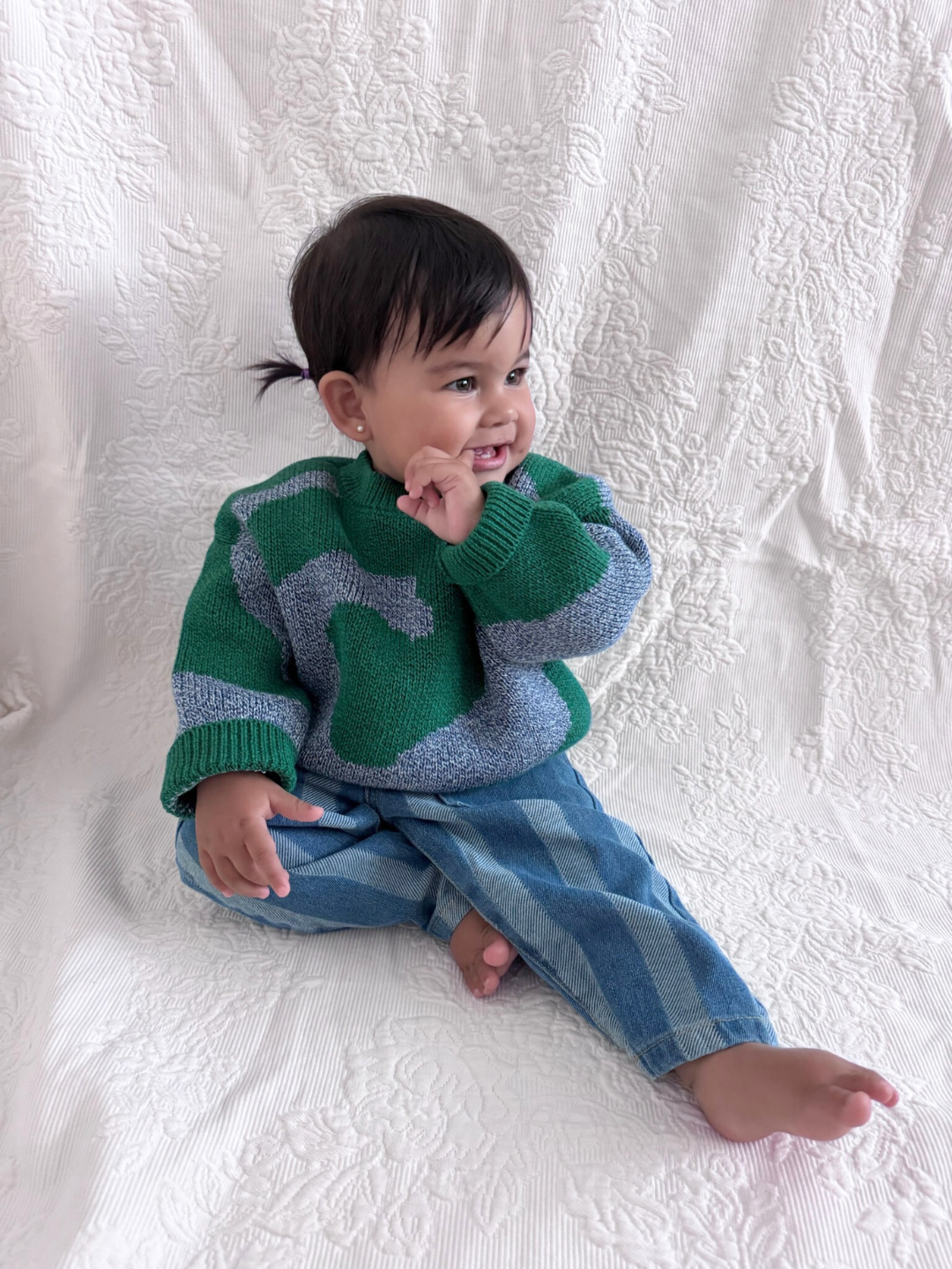 A toddler wearing the SWIRL SWEATER in green and gray with blue plaid pants sits barefoot on textured white fabric, smiling and touching their cheek.