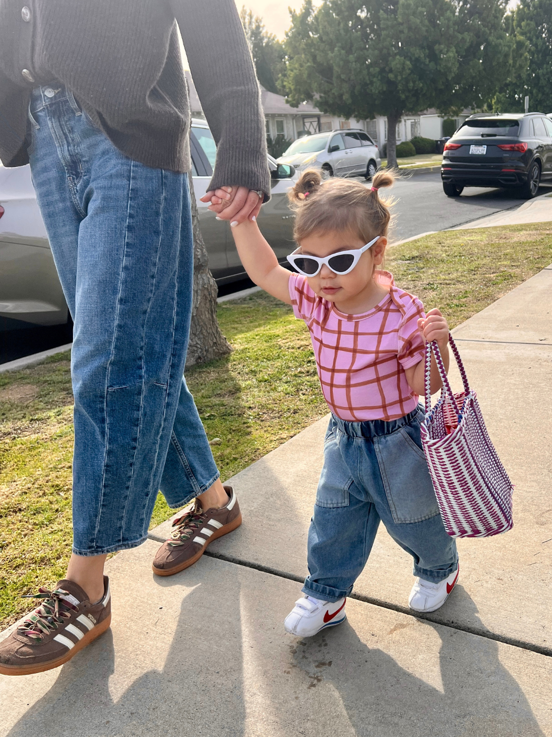 Child holding hands with an adult on a sidewalk, wearing white cat eye sunglasses , baggy blue jeans and a checkered shirt.