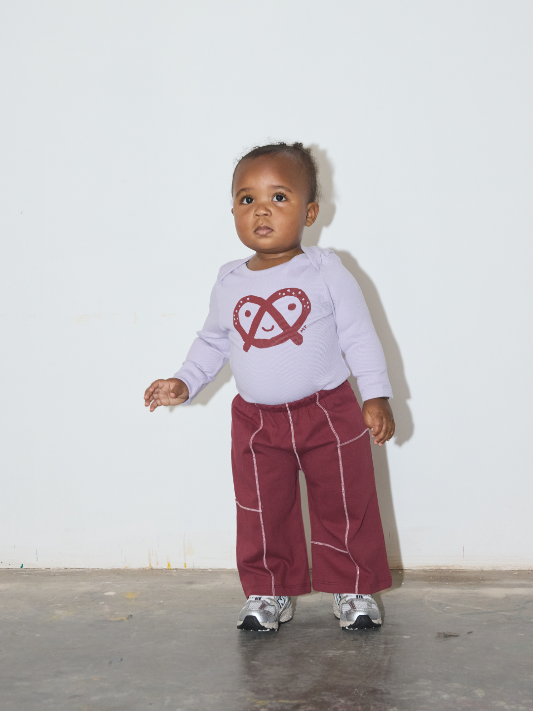 Maroon | A toddler standing against a plain off-white wall wearing a light lavender long-sleeve shirt with a red pretzel graphic and burgundy wide-leg pants with white piping, paired with silver sneakers.