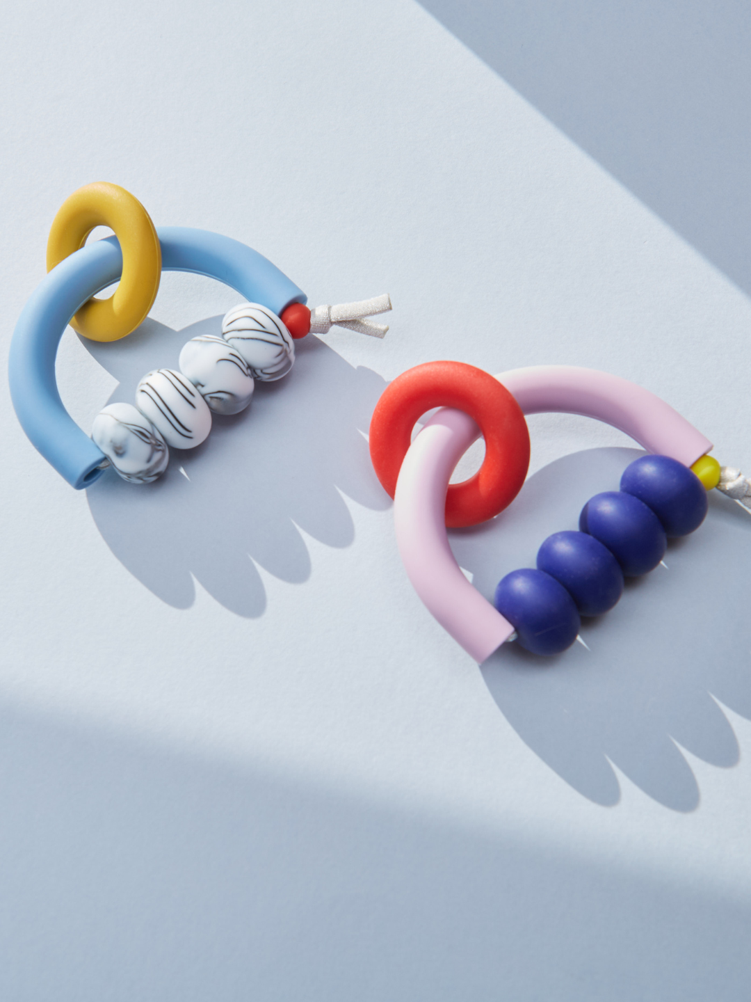 Memphis | Two MT X JM ARCH RING TEETHER silicone baby rings with beads, designed for sensory-friendly teething, are placed on a light blue surface with soft shadows.