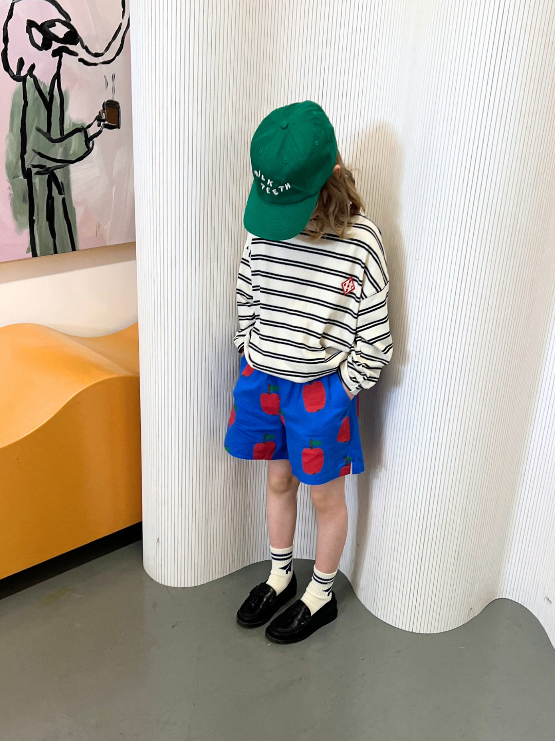 Black & White | A child wearing the PEPPER LONGSLEEVE TEE, a striped cotton top, sports a green cap and blue shorts with red apple prints against a white backdrop. The ensemble from a renowned Korean kids brand is completed with black shoes and white socks.