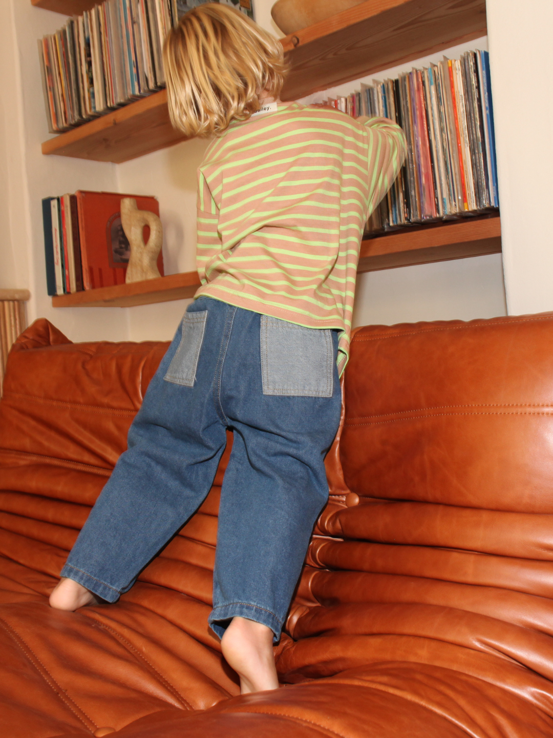 A child reaching for records on a shelf above a brown leather couch wearing the double trouble blue baggy jeans.
