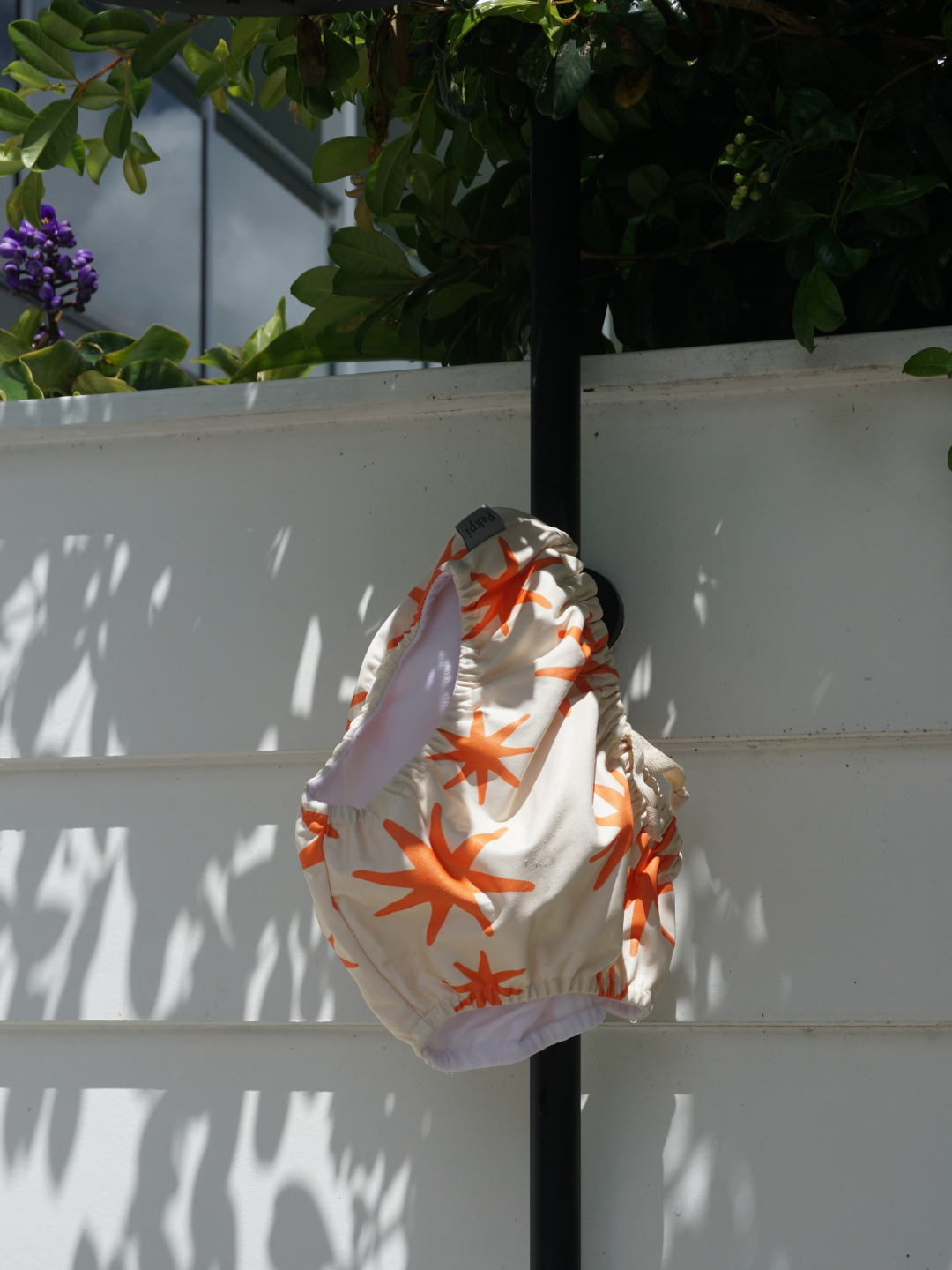 The swim diaper has an elastic waist with a tie and elastic leg holes. The diaper is a cream color with orange stars shapes all over. It is hanging on an outdoor shower in the sunlight.