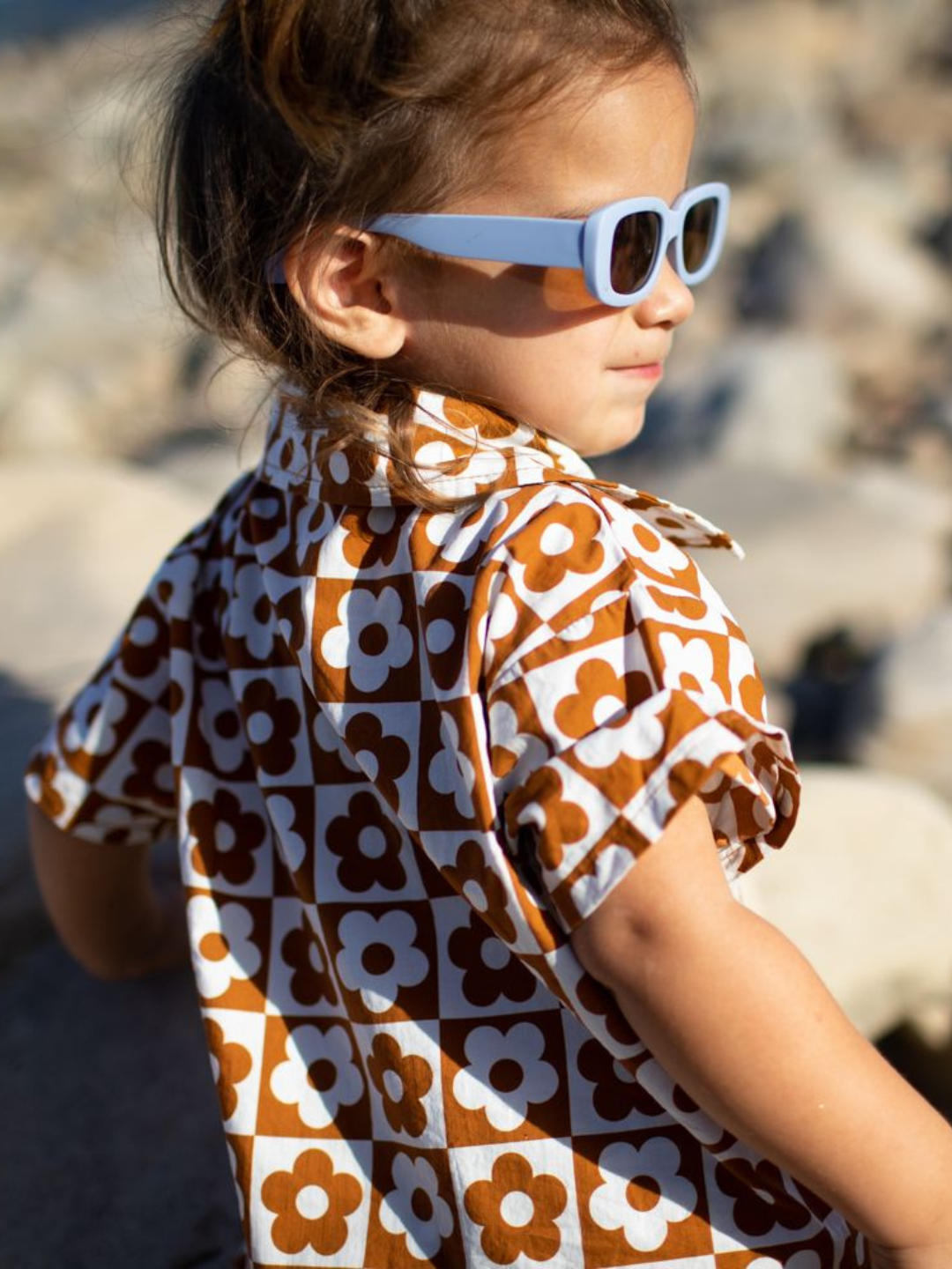 Child in sunglasses showing the shirt of a kids' shirt and shorts set in a checkerboard pattern of terracotta brown and white flowers