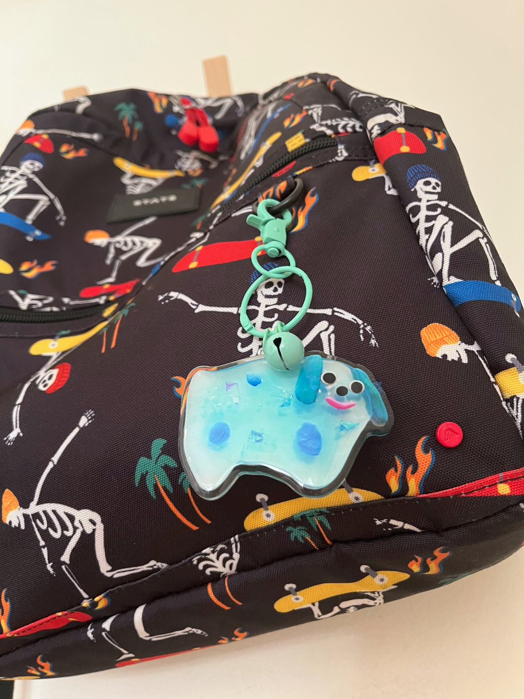 The SOUVENIR KEYCHAIN is a blue, dog-shaped resin charm with spots, a smiley face, bell, and "Thank you" charm. Inspired by Katie Kimmel, all pieces hang from a pale green clasp keyring for cute bag accessories. Shown attached to a kids black backpack in a skateboarding skeletons print.