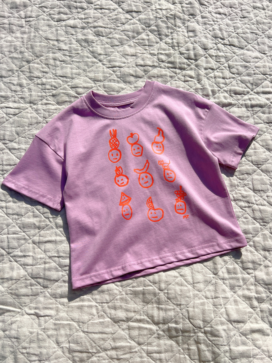 Violet |  A kids' Fruit Face tee in violet is laid flat on a quilt in the sun.
