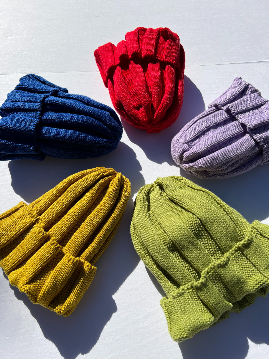 Five knitted kids' beanies laid in a circle: red, lilac, green. mustard, blue