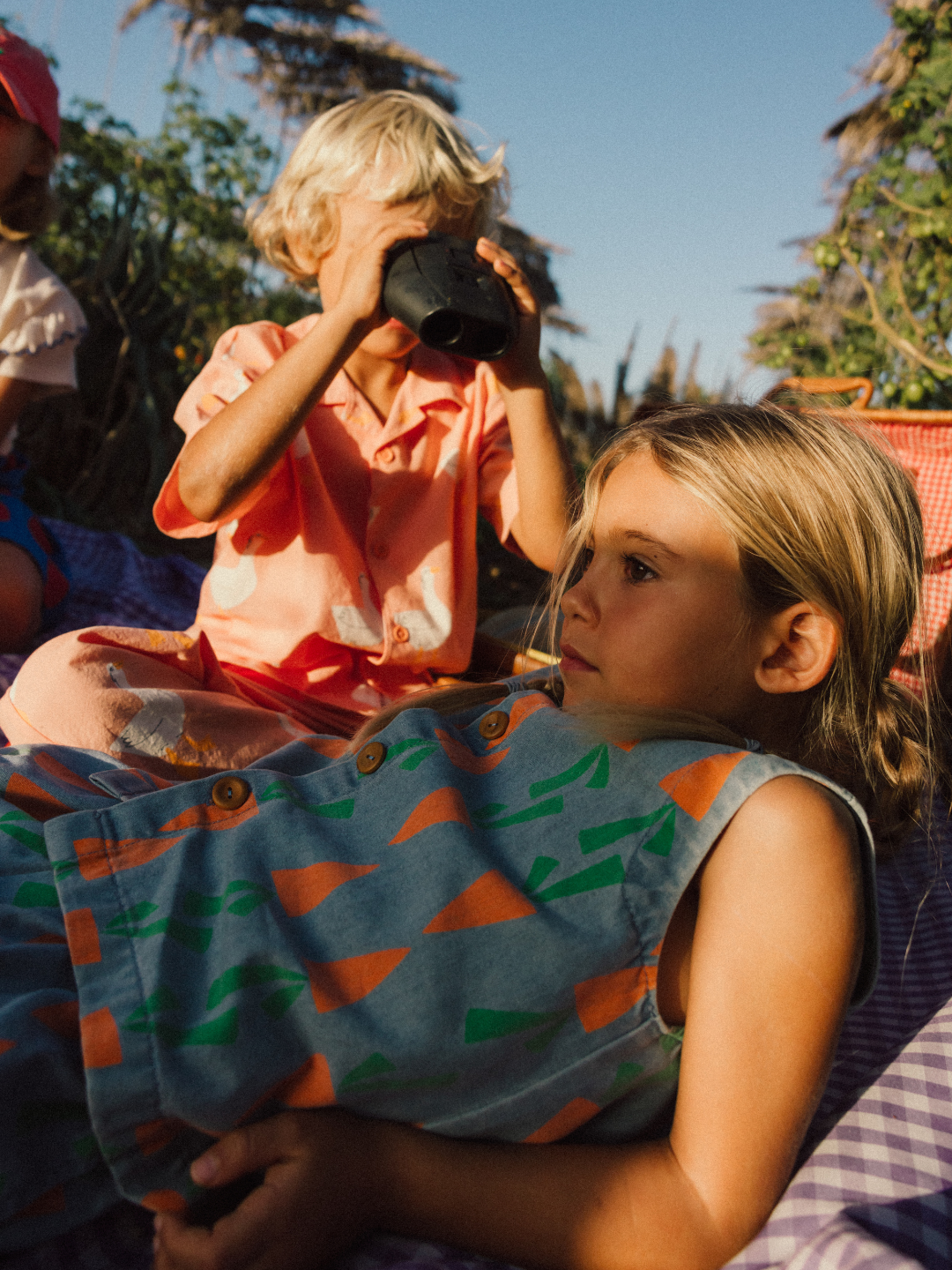 Two children outdoors on a picnic blanket; one reclines, while the other sits with binoculars, both wearing the Carrot Denim Vest.