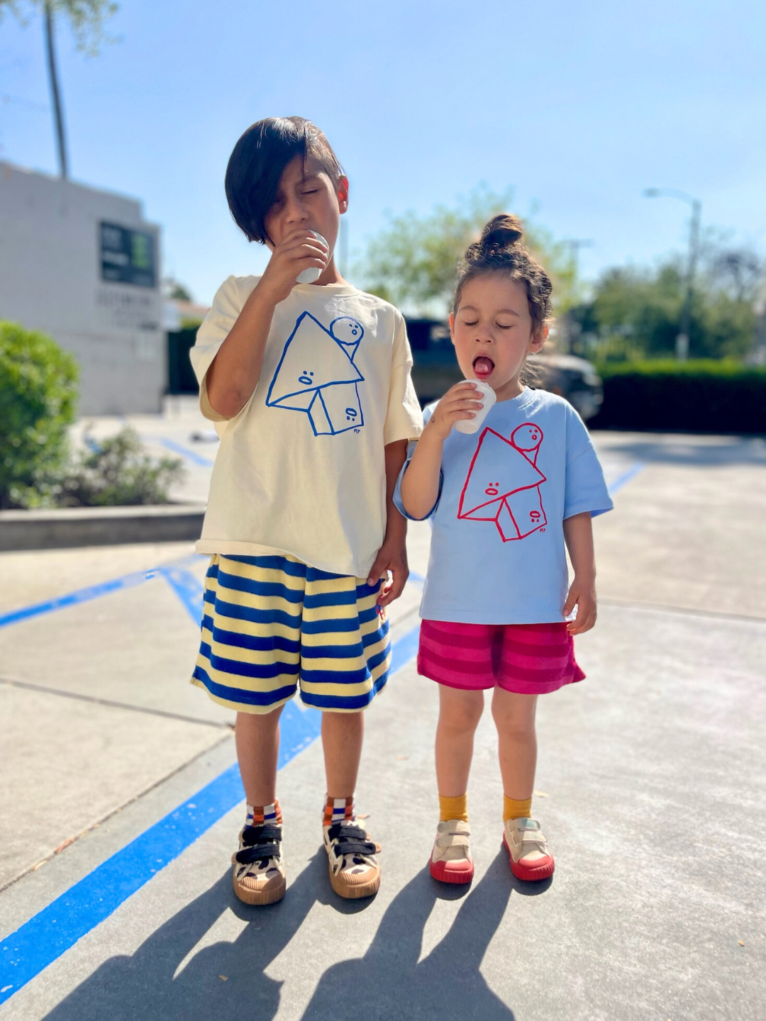 Two kids wearing the Jumble Tee, one in sky blue with red shapes outline with funny faces in the center, and one in the cream shirt with the same design in blue. The kids are wearing striped shirts and eating ice creams, standing in a parking lot with blue lines on the ground, and blue sky behind.