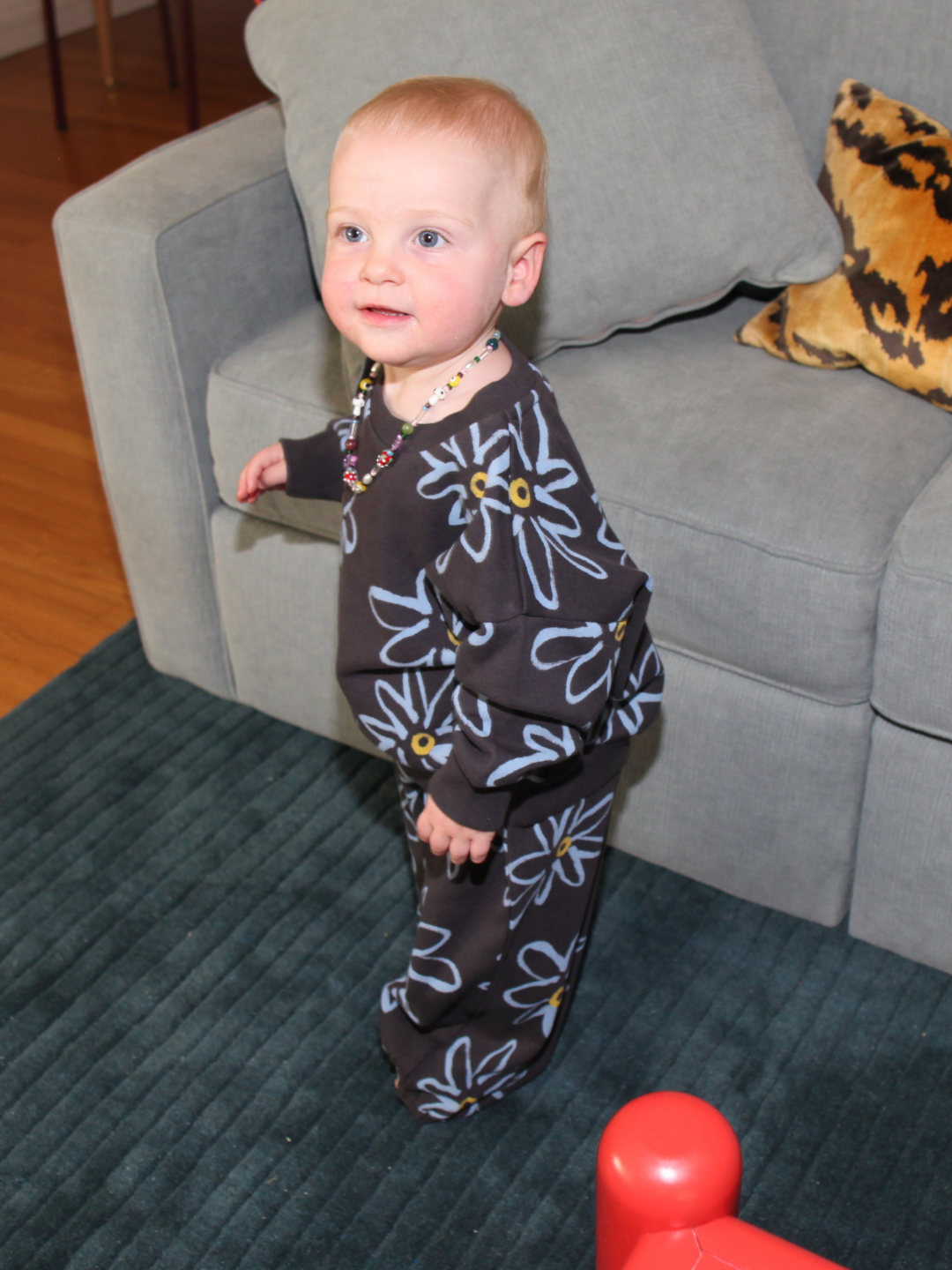 Charcoal | A toddler wearing the CHARLIE SWEAT SET with a painted flower print and a beaded necklace stands beside a gray couch with a pillow, on a blue rug.