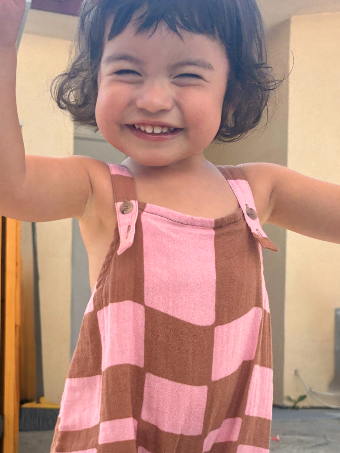 Pink Checkers | Young child smiling with arms raised, wearing MUSLIN OVERALLS, standing outside in front of a beige building.