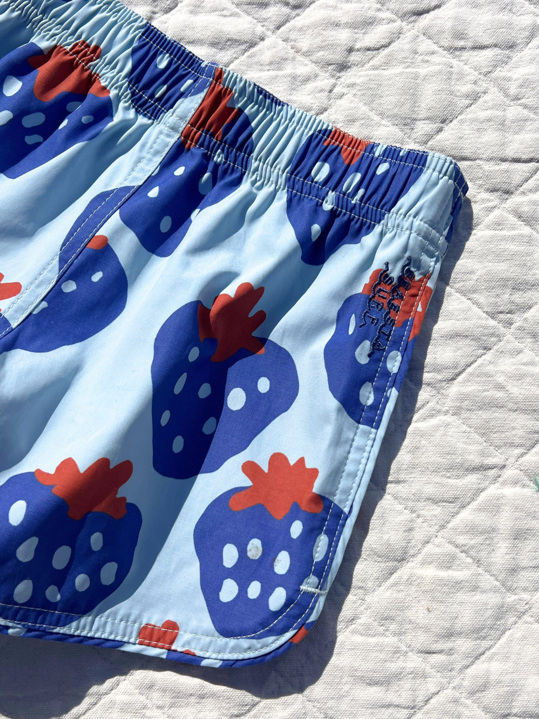 STRAWBERRY BOARDSHORTS feature a playful pattern of strawberries with white dots and red tops on a quilted white background. Made from sustainable materials, these trunks combine retro charm with eco-friendliness.