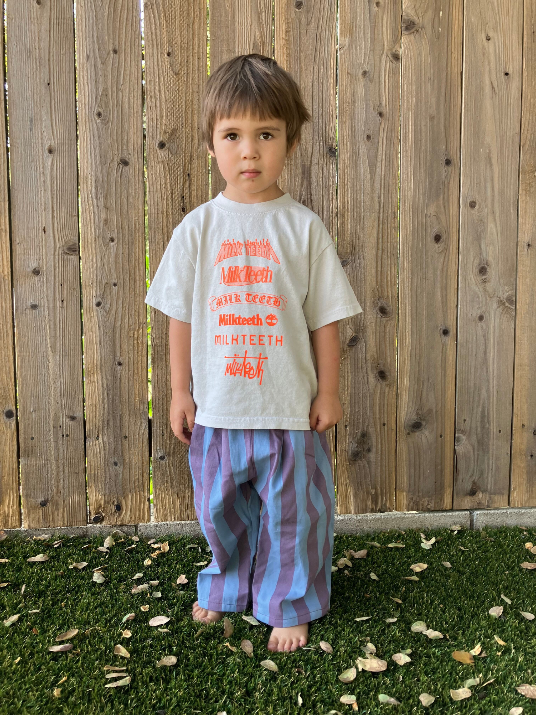 A child wearing a light grey t-shirt with Milk Teeth spelled out in various fonts in orange. He wears striped pants, is barefoot, and stands on grass against a wooden fence.