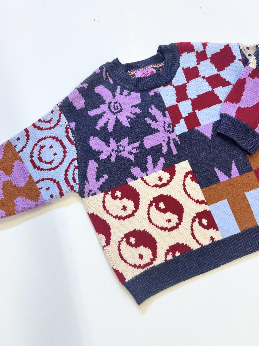 The EVERYTHING SWEATER is a soft knit featuring geometric wavy checkers, smiley faces, animal prints, and yin-yang patterns in red, blue, cream, purple, and brown.