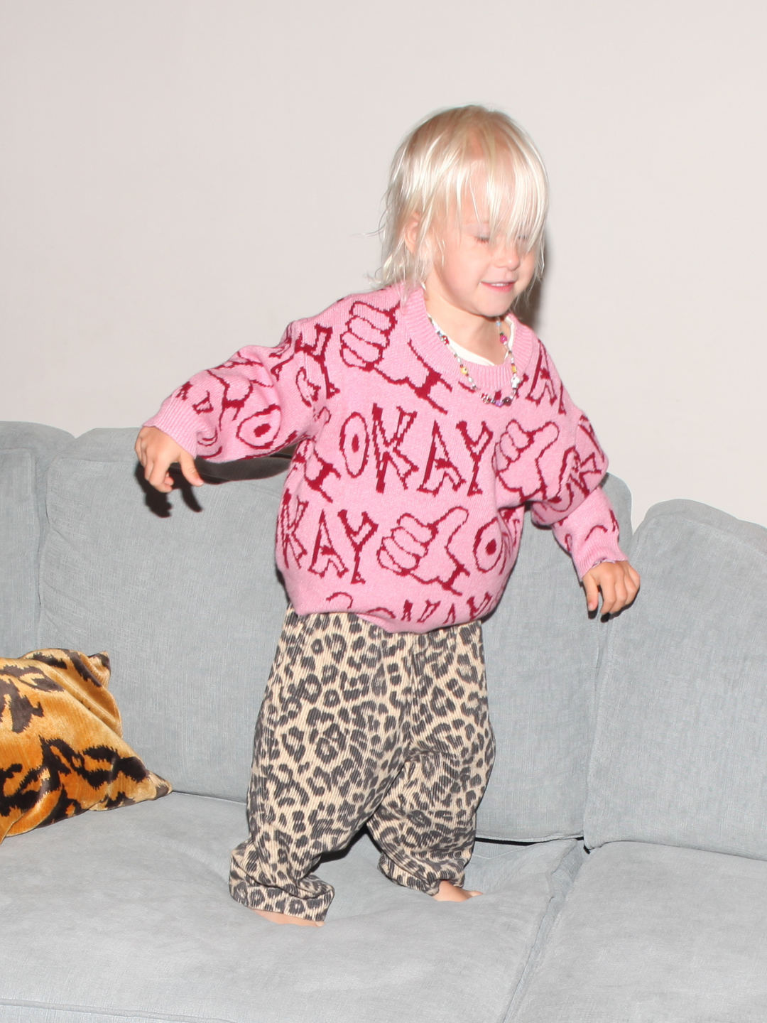Child wearing a pink sweater with text and leopard print pants on a gray couch.