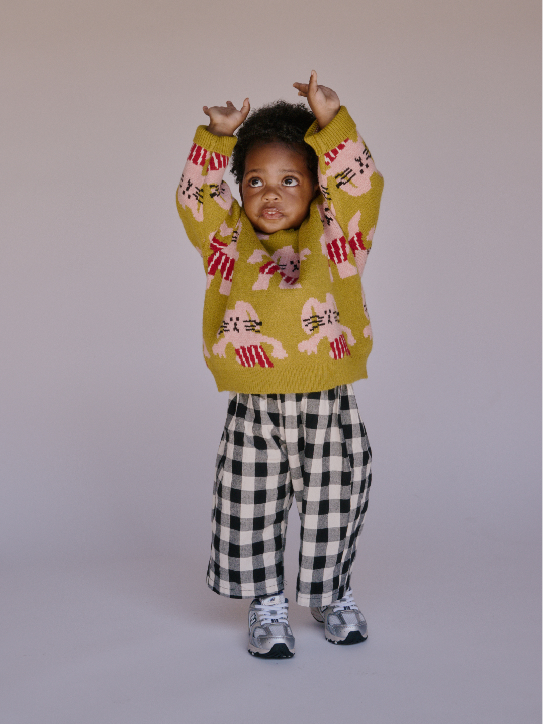 A toddler wearing the MAX SWEATER in yellow with bunny designs, checkered pants, and sneakers stands with arms raised against a plain background.