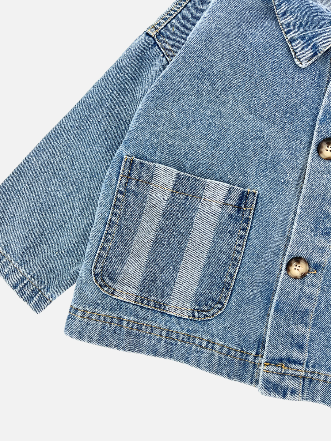 Close-up of the STRIPE POCKET CHORE COAT made from cotton denim, showcasing a chest pocket with striped detail and wood-colored buttons on a light background.