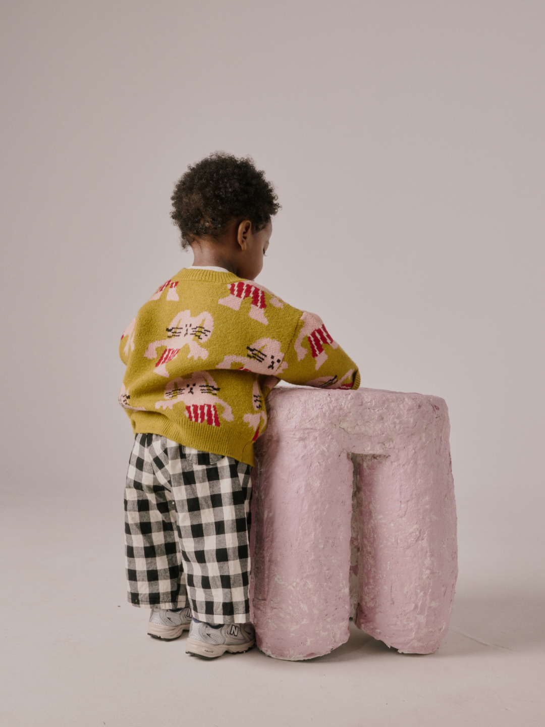 Chartreuse | A young child with curly hair, dressed in the MAX SWEATER featuring bunny designs and checkered pants, stands facing away and leans on a large pink textured object.