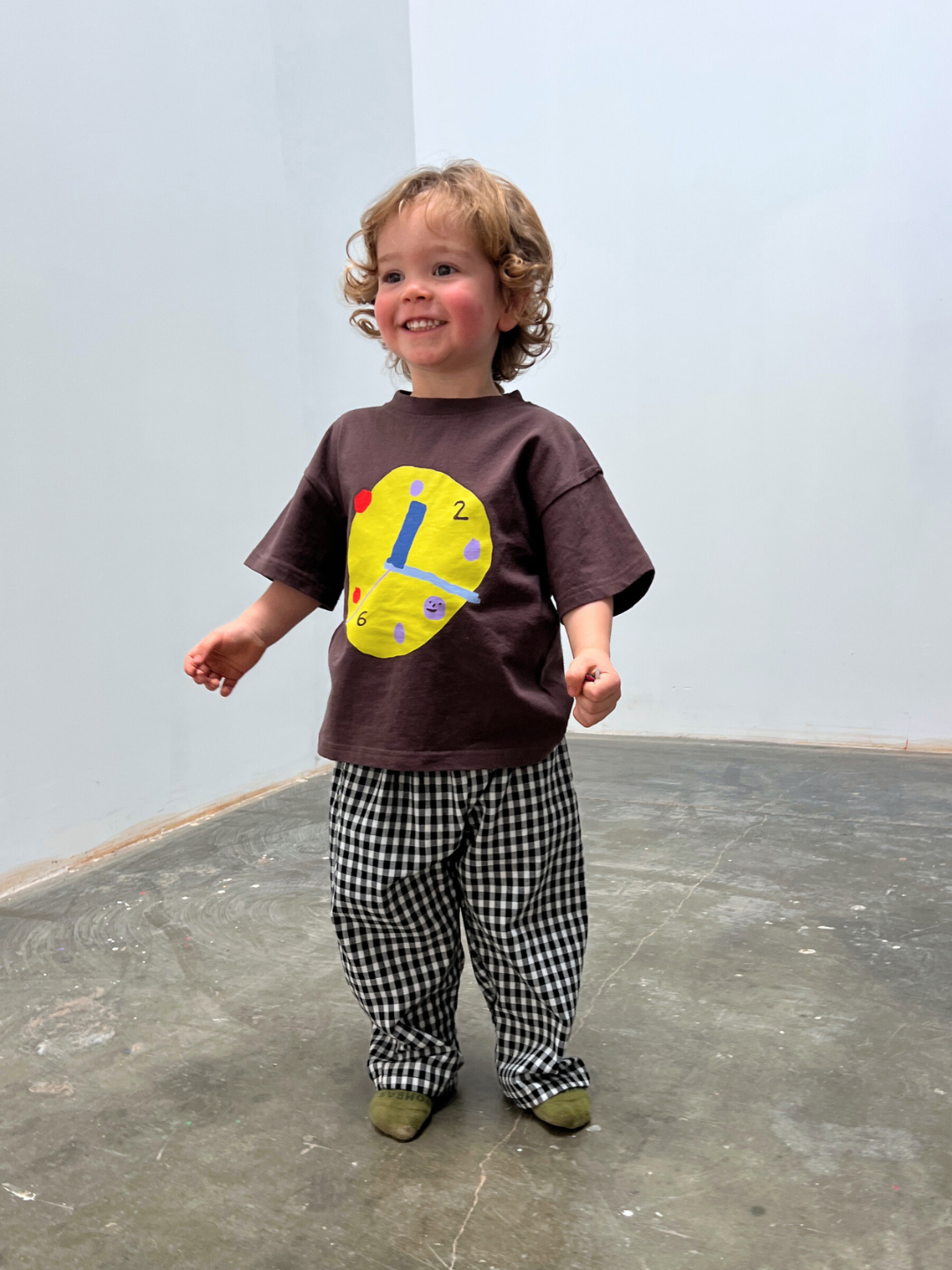 Child wearing a brown shirt with a colorful design and checkered pants on a plain floor.