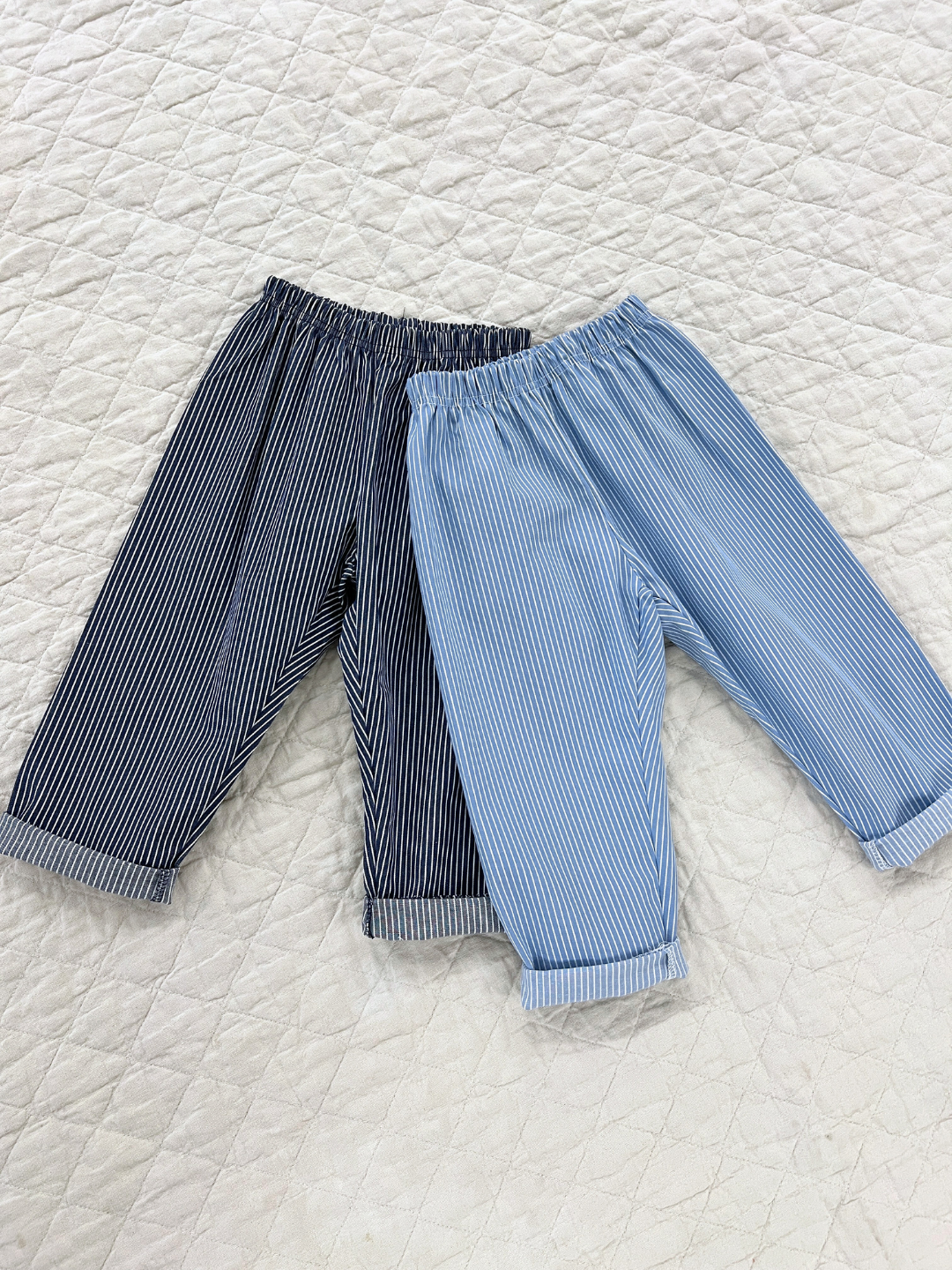 Two pairs of kids blue pants with narrow white vertical stripes, cuffed at the ankle. One is a lighter shade of blue, and one is darker blue. They are laid on a white canvas quilt.