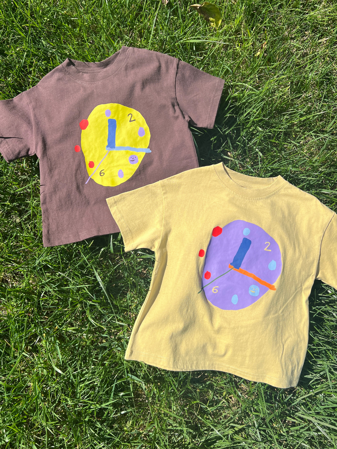 Two children's t-shirts with clock designs on a grassy background