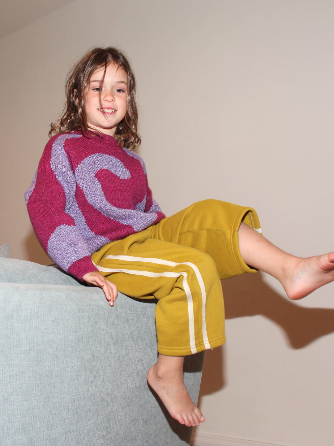 Pink/Purple | Child wearing a pink sweater with a purple design and yellow pants with white stripes, sitting on a gray couch.