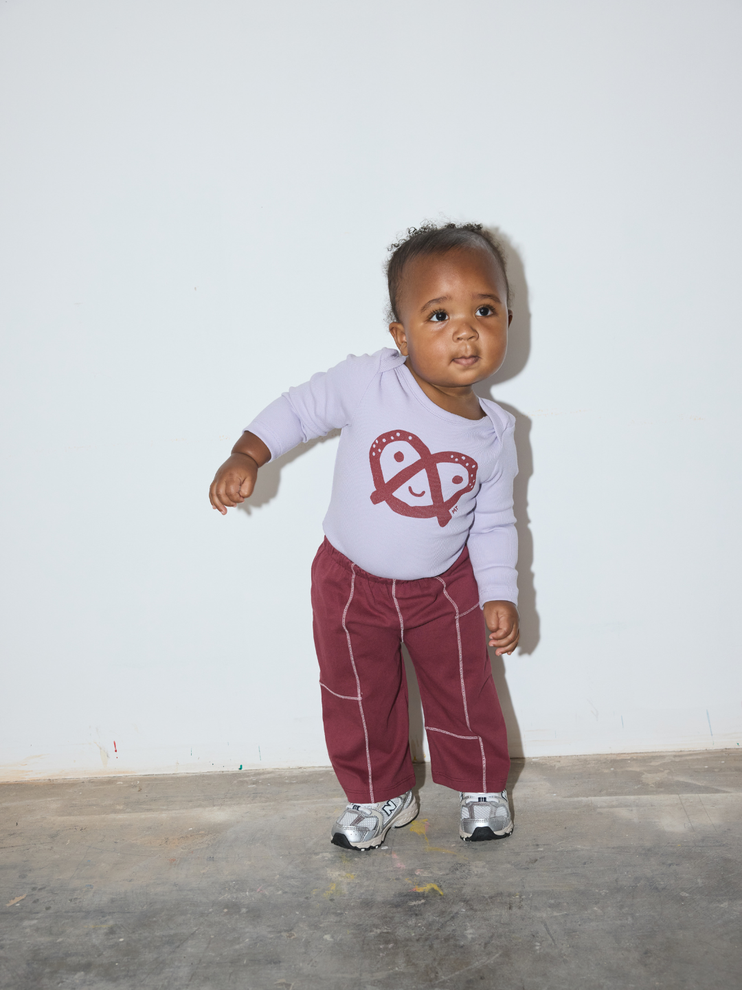 A toddler standing against a plain white wall wearing a light lavender long-sleeve top with a red graphic print and burgundy pants with white piping.