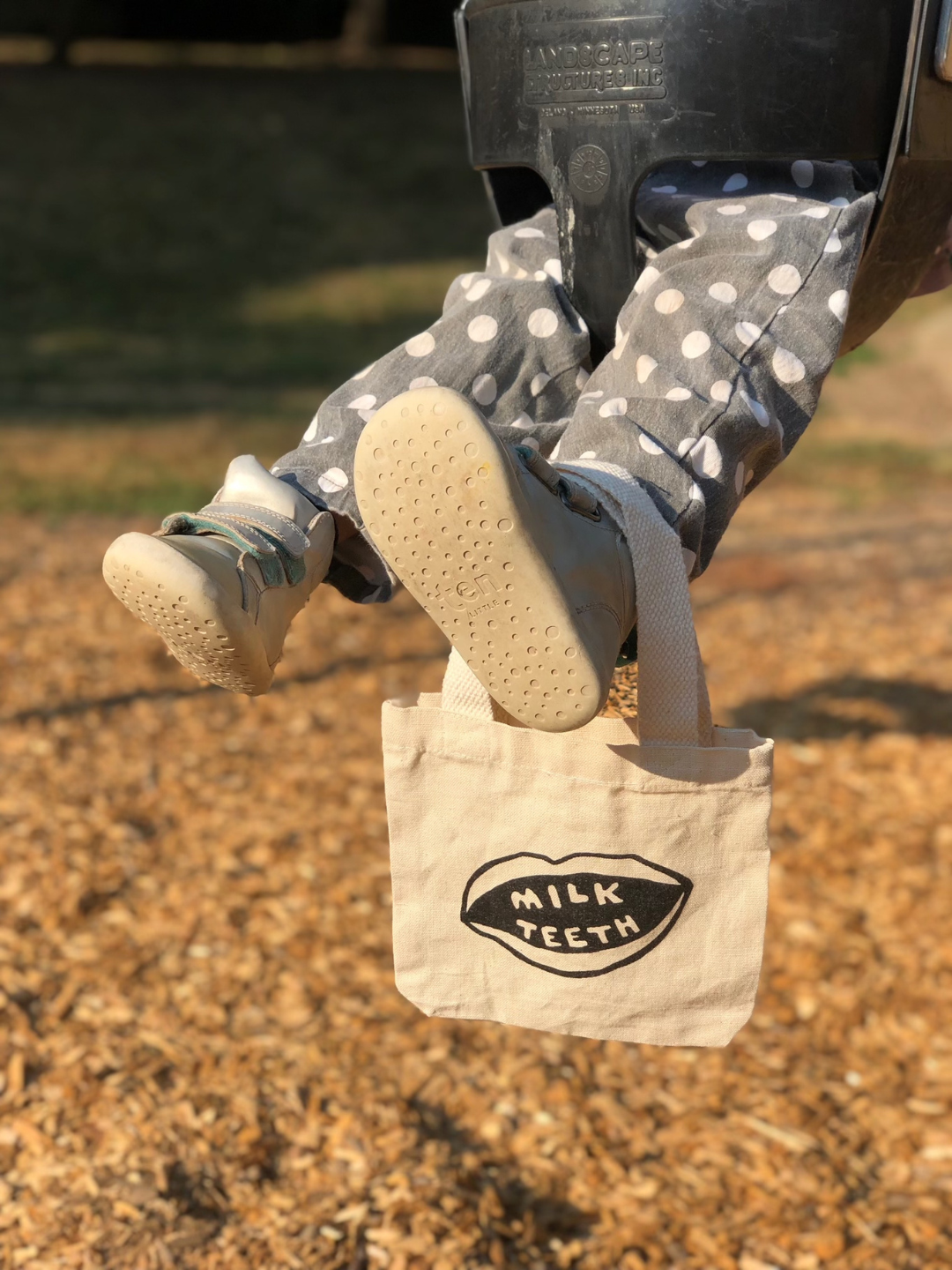 A Milk Teeth mini tote is hanging off the child's foot as they are swinging. 