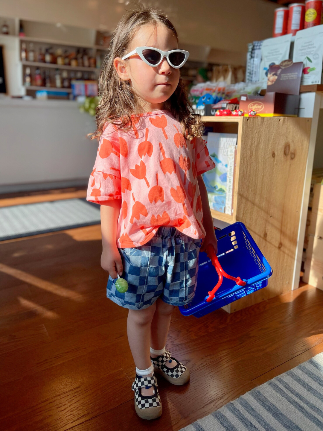 Child with long dark hair wearing the tulip tee with blue denim checkered shorts, white cat eye sunglasses, black and white checkered mary jane sneakers and white ankle socks. She is holding a blue shopping basket inside a grocery store.