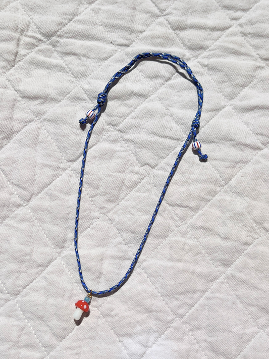 Image of The FUNGI NECKLACE by Meyous jewelry is a handmade, genderless blue cord necklace with an adjustable fit and a red and white glass mushroom charm, shown on quilted white fabric—ideal for any style.