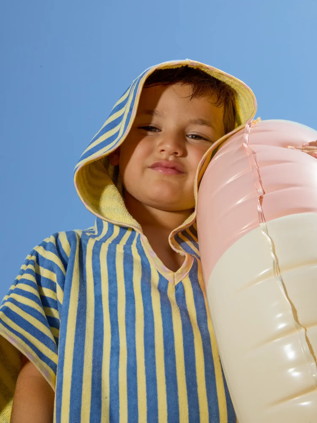 A child in the STRIPED PONCHO holds a large inflatable ice cream cone under a clear blue sky.