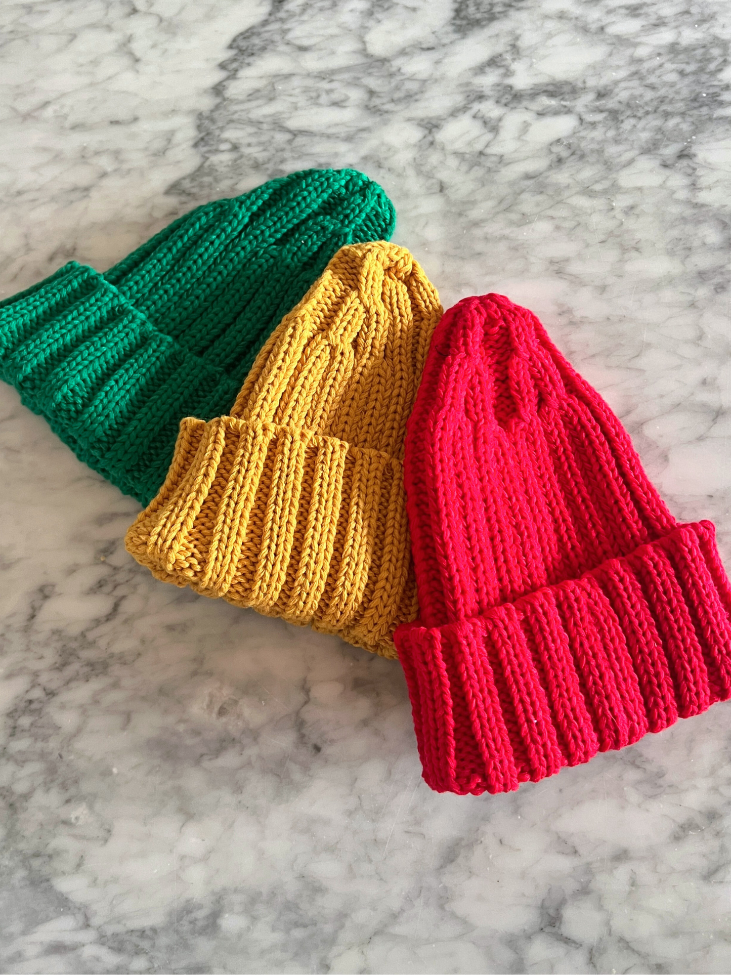 Three CHUNKY RIB KNIT BEANIE hats in green, yellow, and red sit side by side on a gray marble surface, each with an adjustable cuff for a perfect fit.