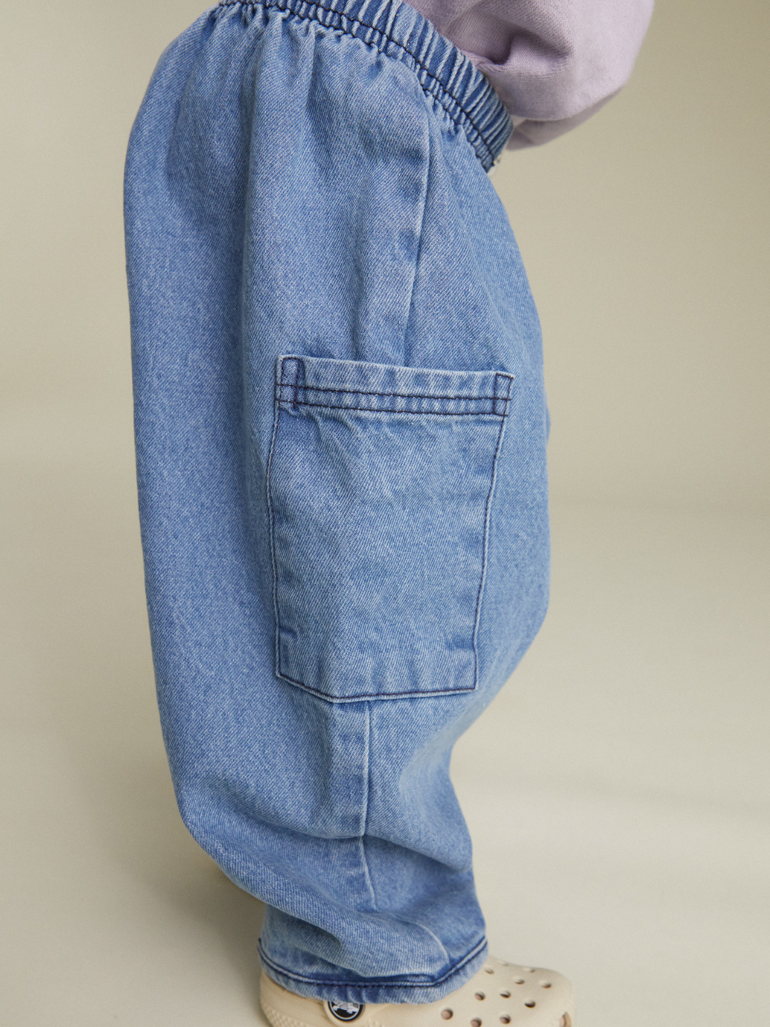 A close-up side view of a person wearing EMERSON DENIM BABY TROUSER—unisex organic cotton denim pants with an elastic waistband, large side pocket, and light-colored shoes.