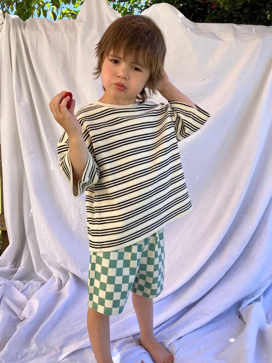 Teal | A child wearing the kids Frankie Short in Teal & Ivory check, paired with a boxy cream tshirt with fine black stripes. He is eating a strawberry and standing on a white cloth backdrop, with trees in the background.