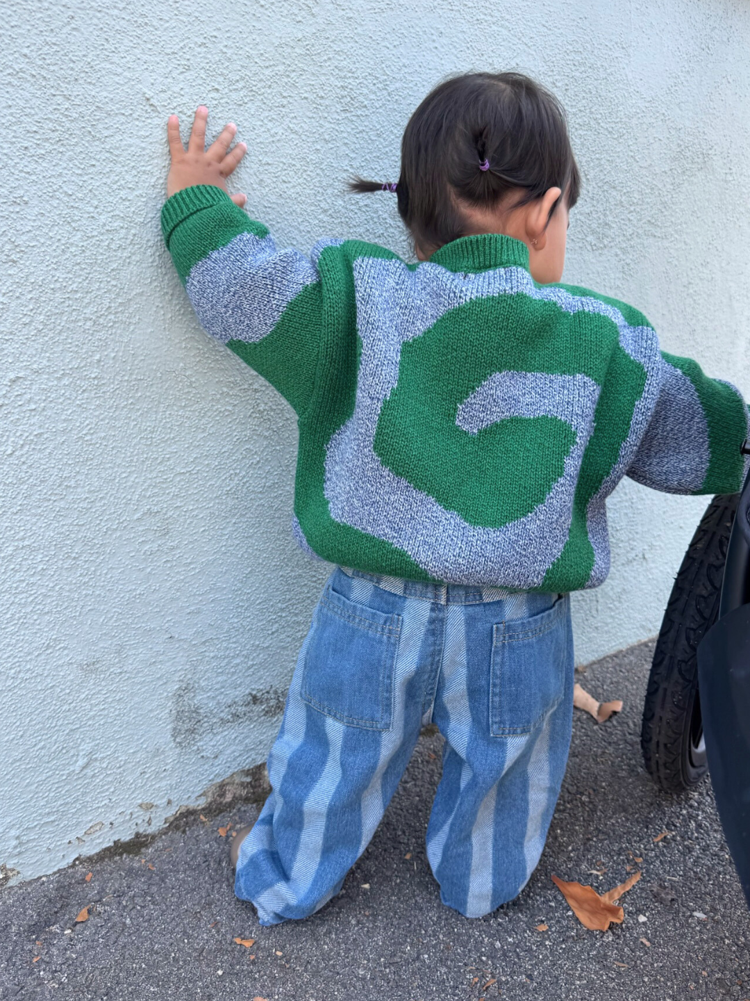 Green/Blue | A small child with two pigtails faces a wall, wearing the SWIRL SWEATER in green and gray and blue striped jeans.
