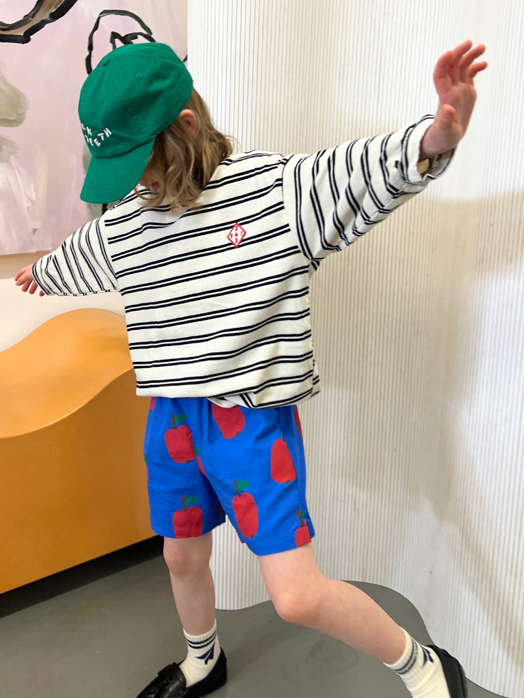 A child balances indoors with arms outstretched, wearing a PEPPER LONGSLEEVE TEE with stripes, paired with blue shorts adorned with red apples and a green cap. The ensemble showcases the boxy silhouette style emblematic of trendy Korean kids' fashion.