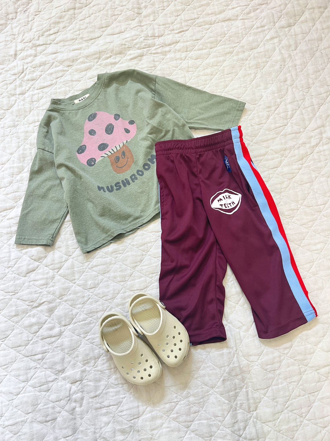 Green | A children's outfit on a quilted surface includes a green Mushroom Long Sleeve Tee, maroon pants with blue and red stripes, and cream slip-on shoes.