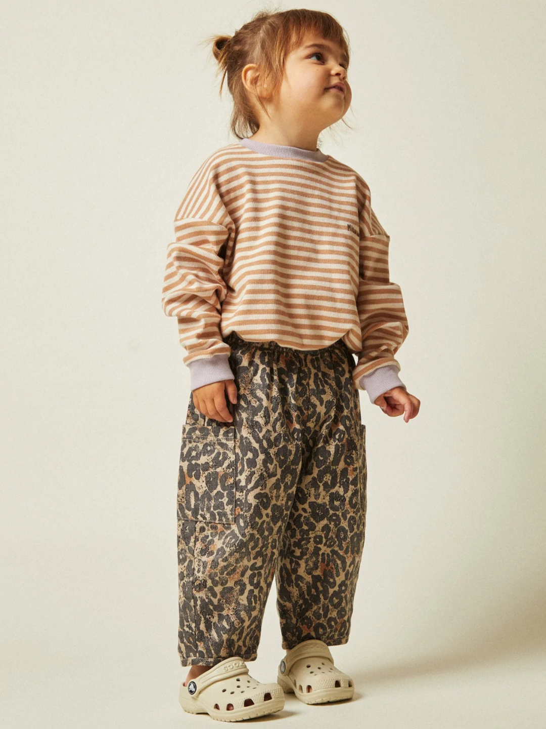 A young child stands against a plain background wearing beige and white stripes with EMERSON LEOPARD TROUSER NEW FIT, made from organic cotton, paired with light slip-on shoes.