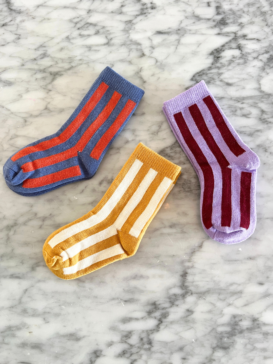 Violet | Three pairs of STRIPE SOCK on a marble surface: one pair is blue with red stripes, one pair is yellow with white stripes, and one pair is lavender with maroon stripes. Made from stretchy cotton, these comfy socks are perfect for any occasion.