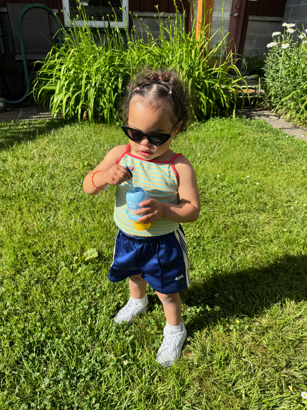 Child in black cats eye sunglasses and blue shorts standing on grass holding a container of bubbles.