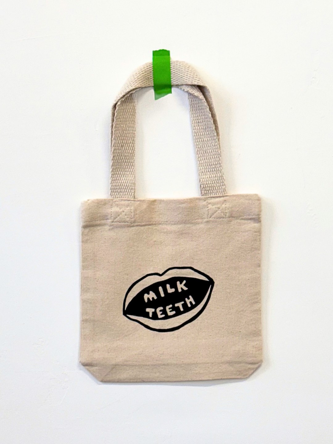 Milk Teeth mini tote front view, hanging on the wall. Cotton tote with Milk Teeth mouth logo in the center.