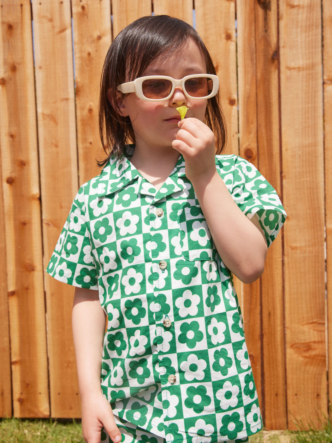 Child in sunglasses showing the shirt of a kids' short set in a checkerboard pattern of green and white flowers